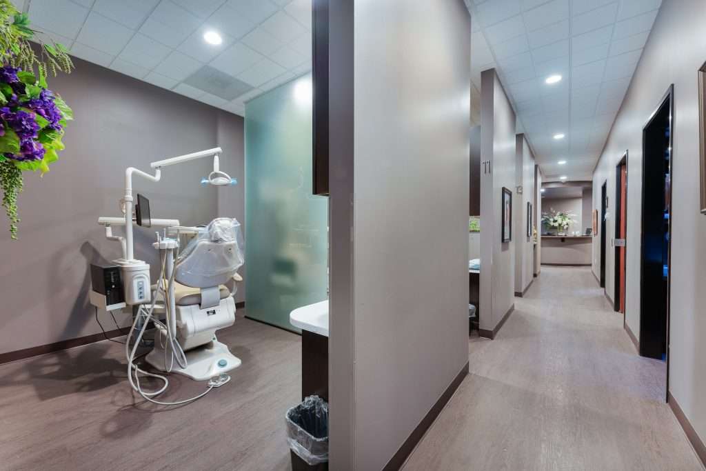 Clean modern operatory with chair and overhead monitor beside a bright hallway at Sacramento Dentistry Group